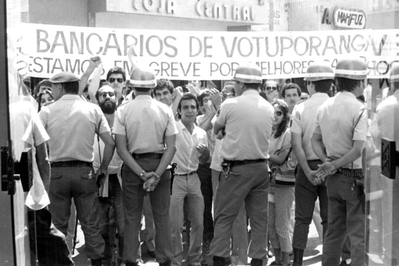 Greve dos Bancários  Nos anos 80, pouco depois da instalação do Sindicato dos Bancários de Votuporanga, houve uma greve da categoria e o pessoal procurou mostrar serviço. Esta foto foi na rua Amazonas defronte a um estabelecimento bancário. Entre os manifestantes, pode ser identificado o Achiles, que tempo depois assumir a presidência da entidade. A foto é do arquivo do jornal A Cidade.
