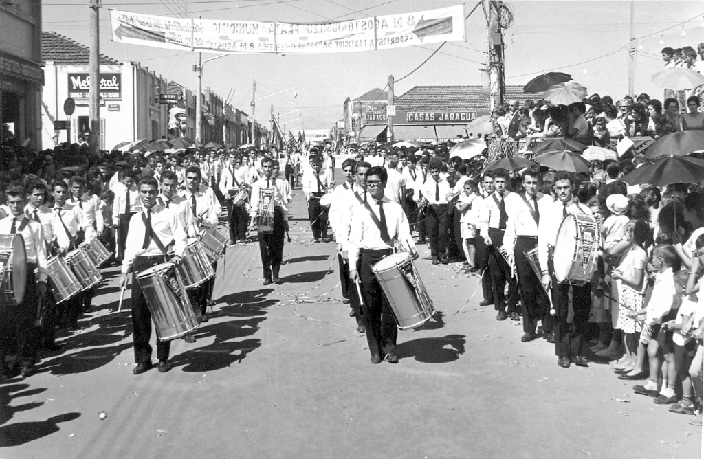 A fanfarra Ginásio Estadual e Escola Normal “Dr. José Manoel Lobo”, no desfile cívico de 1960. Da esquerda para a direita: João De Haro, Maurício Silvério Gomes, Nagata, João Gualberto Rosa, Dioraci, Aldeziro Padovani, Manoel Braolho e Oswaldo Padovez. No palanque dá para ser identificado o prefeito Hernani de Matos Nabuco, e ao fundo o prédio de Casas Jaraguá, na esquina da Concha Acústica. A foto é José Zucarellli.