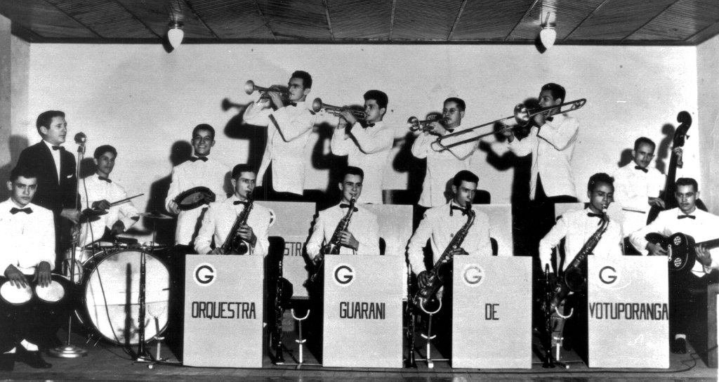 Nos anos 50 e 60, tempo em que os bailes de gala eram animados ao som das grandes orquestras, Votuporanga exportava o sucesso da orquestra Guarani, que se apresentava por este Brasil a fora. Alguns dos seus músicos ainda continuam na ativa, mas a época de ouro das grandes orquestras ficou na saudade. Para recordar, eis aí a composição da Orquestra Guarani. Da esquerda para a direita aparecem na segunda fila tocando pistom: Leopoldo, Ademar e Oliveira. No trombone Vadão e no baixo Jair. Na primeira fila, o bongô com João Castilho, atrás dele o cantor Valdir, bateria Aloísio; pandeiro Lourival, e fila de sax Leonel Riva, Dubair, Rômulo e Tesourinha. Finalmente, o violão de Riva. Esta foto pertence a João Castilho, nosso leitor, residente em Guapiaçu-SP.