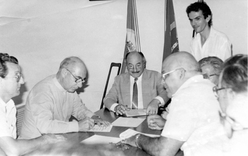 Ano 85 - O Bispo Diocesano D. José de Aquino Pereira, aparece nesta foto assinando documentos relativo a sessão de uma área por parte da Prefeitura à Igreja. Aparecem na foto, a partir da esquerda: Antonio Domingo Barbon, Bispo D. José , o então prefeito Mário Pozzobon, o assessor Luciano Viana (de pé) e o vereador Meidão, entre outros.
