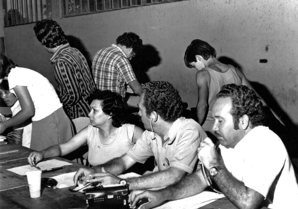 Em ação a equipe da Rádio Clube AM na cobertura da apuração dos votos na eleição de 1976. Naquele pleito eleitoral o dentista João Antonio Nucci elegeu-se prefeito pela primeira vez. O radialista Aguinaldo de Oliveira (à direita com o microfone) foi eleito o vereador mais votado com 768 votos pela Arena. Na esquerda, o técnico de som Shoyo e na mesa o casal de radialistas Luiz Rivoiro e Oneide Aparecida Lourenço Rivoiro. Atrás, de costas, Edélcio Martins e Celso Fiorentino. A foto pertence ao álbum de Aguinaldo de Oliveira