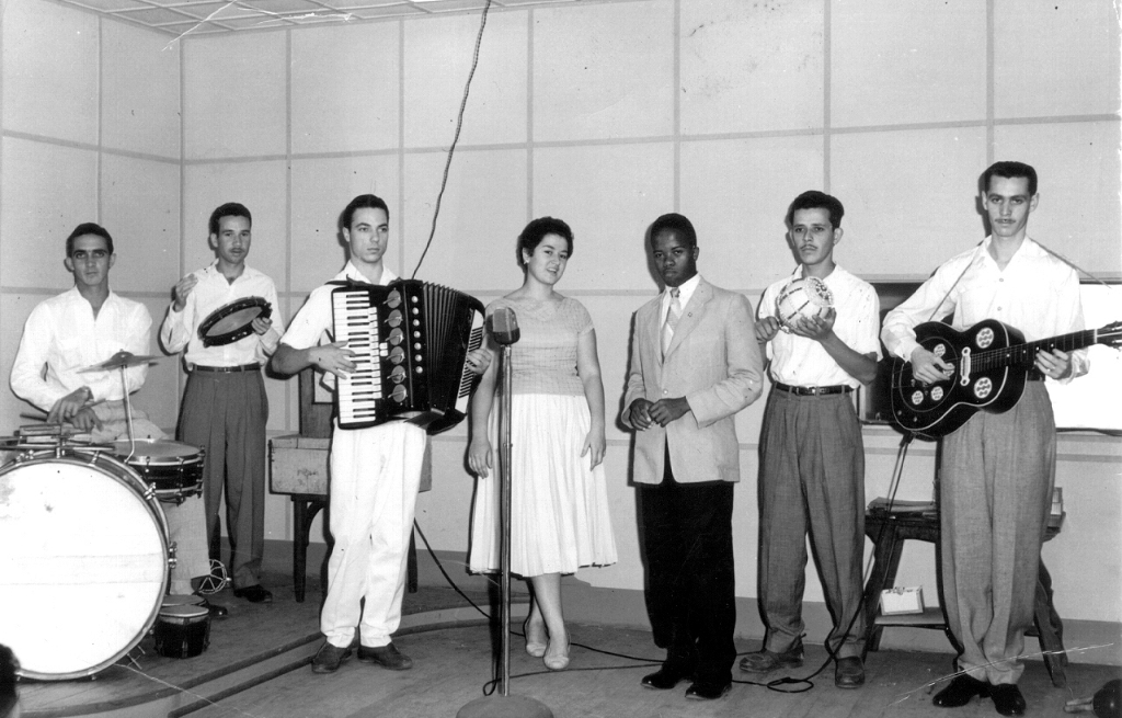 Era do Rádio. O ano é de 1956. Ano de inauguração da Z.Y.R. 200 - Rádio Clube de Votuporanga. Programa de auditório e de calouros aos domingos de manhã. Na foto, da esquerda para direita: Darcy Secchini (bateria); Aguinaldo de Oliveira (pandeiro); Armando Domenicis (acordeão); Maria do Carmo (cantora); Geraldo de Almeida (cantor); Rames Cury (afoxê) e Amilar Riva (violão). Apresentavam-se ainda, Claudenice Públio - a bonequinha loira; Abilio Ribeiro - o Abilio Baião, como intérpretes preferidos - destacando ainda, as irmãs Esmeralda, Marivalda e Ivone Góes de vozes maravilhosas. Aguinaldo de Oliveira e Rames Cury aparecem na foto para dar ênfase ao conjunto, pois gostavam, mas não eram músicos e sim locutores de rádio. A foto é do acervo de Amilar Riva