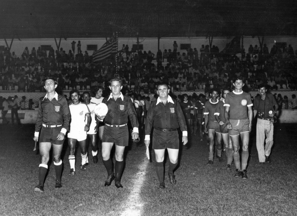 Decisão do Campeonato Amador Regional na tarde 23 de novembro de 1975. Local: Estádio Plínio Marin. O momento da entrada em campo dos times finalistas: Riolândia e Cardoso. Na frente, o trio de arbitragem da Liga Votuporanguense de Futebol. Na esquerda, o bandeira amarela Sebastião Batista da Silva Filho, ao centro, o árbitro Roberto Bianchini, e na direita, o bandeira vermelha Roberto Popoli. O jogo foi decidido nos pênaltis e deu Cardoso. 