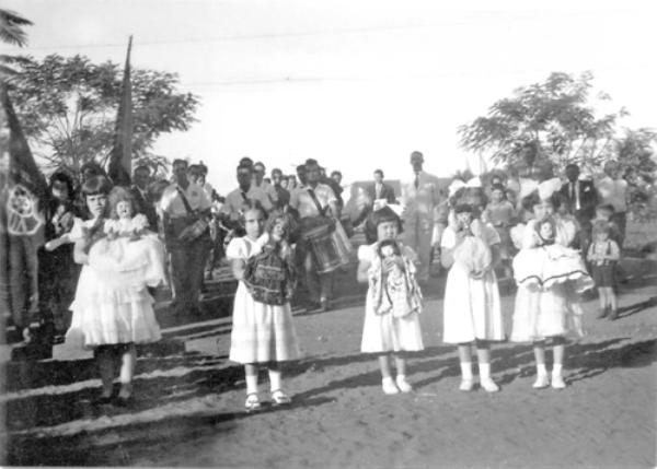 Desfile das Bonecas - Na década de 50, a quermesse em prol da construção da Santa Casa tinha como ponto alto o Leilão das Bonecas. As bonecas especialmente confeccionadas para o leilão representavam as colonias já radicadas em Votuporanga. Nesta foto, o professor Laonte comandava a fanfarra (ao fundo) que chamava a população para participar das noitadas de quermesse que aconteciam no largo da Matriz. Em primeiro plano, aparecem as crianças conduzindo as bonecas das colônias. Da esquerda para a direita: primeira boneca era da Santa Casa; a segunda Portuguesa; a terceira Japonesa, a quarta Turca (Sírio-Libanês) ; e a quinta italiana.A foto pertence ao arquivo pessoal de Joaquim Marques.