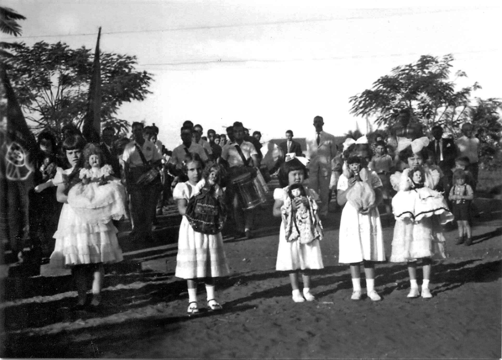 Na década de 50, a quermesse em prol da construção da Santa Casa tinha como ponto alto o Leilão das Bonecas. As bonecas especialmente confeccionadas para o leilão representavam as colonias já radicadas em Votuporanga. Nesta foto, o professor Laonte comandava a fanfarra (ao fundo), que chamava a população para participar das noitadas de quermesse, que aconteciam no largo da Matriz. Em primeiro plano, aparecem as crianças conduzindo as bonecas das colônias. Da esquerda para a direita: primeira boneca era da Santa Casa; a segunda Portuguesa; a terceira Japonesa, a quarta Turca (Sírio-Libanês); e a quinta Italiana. A festa sempre foi um grande sucesso