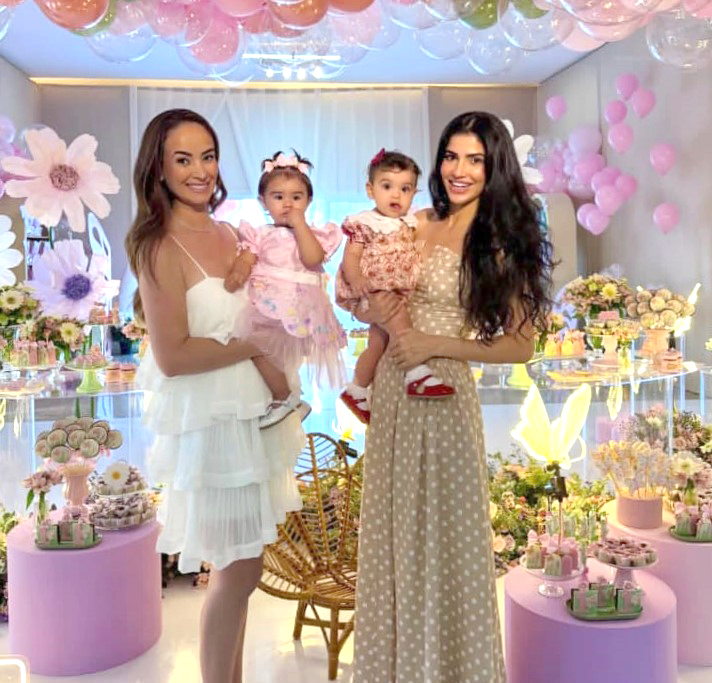 Ana Carolina Guimarães Scamatti com a princesinha Ana Luísa Scamatti ao colo, acompanhada pela Dra. Josiane Frederico com a filha Helena Costa Frederico. Ao fundo, um encantador cenário cor-de-rosa, com um delicado jardim repleto de borboletas, tema escolhido para celebrar o 1º aninho da pequena Ana Luísa, filha de Ana Carolina e Luiz Scamatti. Felicidades, Ana Luísa! Que sua vida seja sempre iluminada, repleta de alegria, amor e que você siga crescendo com muita saúde e rodeada de carinho.