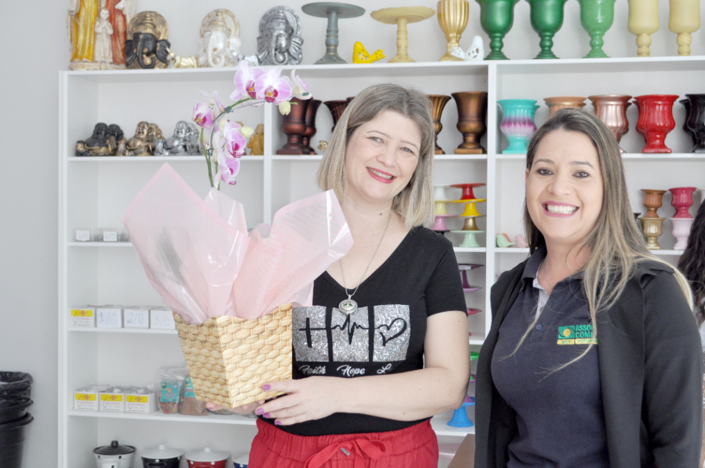 A proprietária da loja “Hader Magazine” sendo gentilmente presenteada com flores por Jordana, representando a ACV, que tem por costume presentear todo comerciante que inaugura uma loja na cidade, para dar as boas vinda e boa sorte